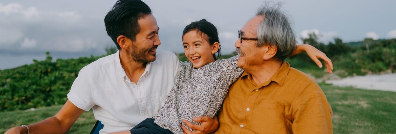 A happy child sits between her father and grandfather on a grassy area with a body of water in the background