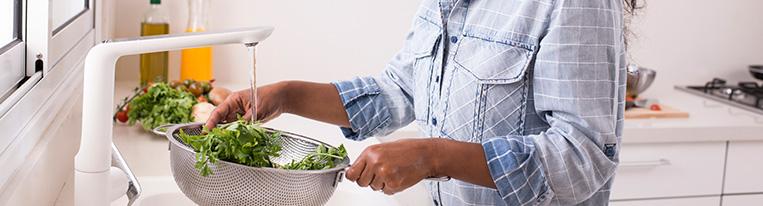 A woman rinses parsley in a colander at her kitchen sink.