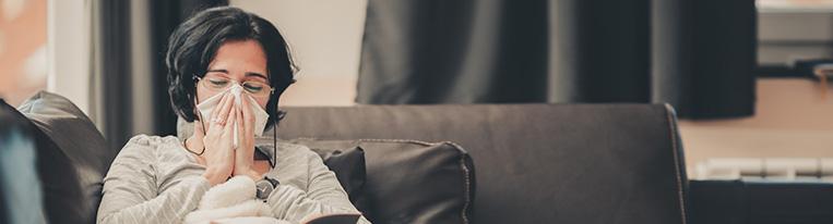 A woman lying on a couch under a blanket blows her nose with a tissue while reading a book that sits in her lap.