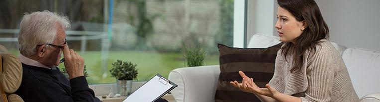 A young woman talks to a therapist, who sits across from her and holds a pen and clipboard.