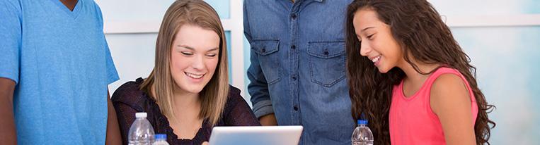 Four adolescents gathered around a table look at a tablet together.