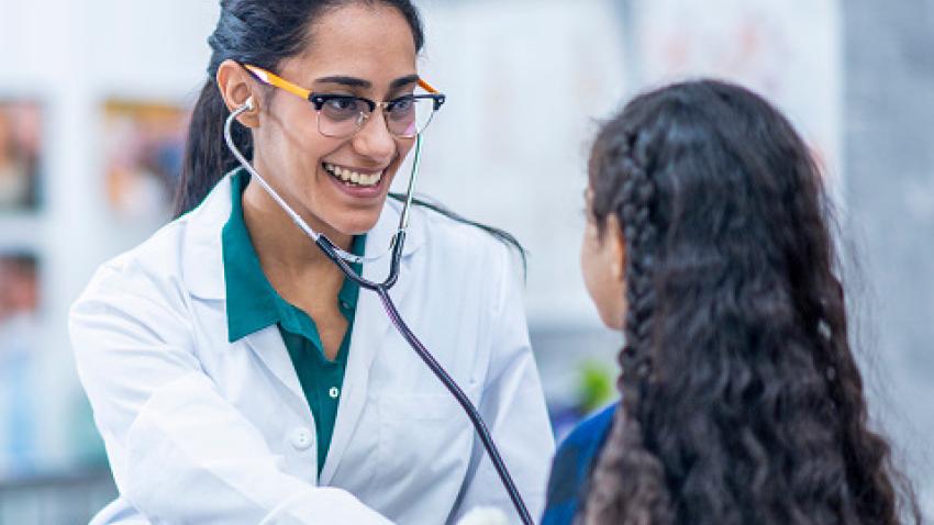 A doctor with long dark hair in a ponytail, glasses, and a white lab coat smiles and checks a child’s  vital signs using a stethoscope.