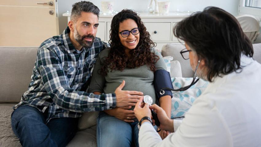 A pregnant woman and partner sit on a couch while a clinician wearing white lab coat checks the pregnant person’s blood pressure.